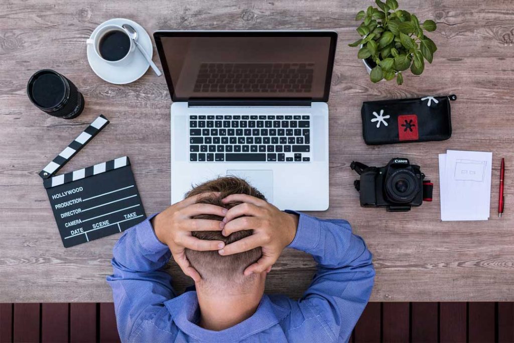 Das Bild zeigt einen verzweifelten Blogger an seinem Schreibtisch mit einem Notebook, Kaffee, einer Filmklappe, Digitalkamera und einem Notizblock.